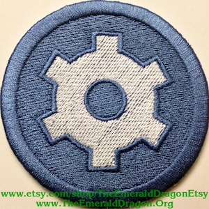 May include: Embroidered patch featuring a gear design in light gray and navy blue. The gear is centered within a navy blue circular border. The patch is likely for clothing or accessories.