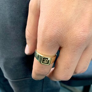 May include: A gold ring with a square-shaped setting featuring a black letter 'D' and a small diamond.