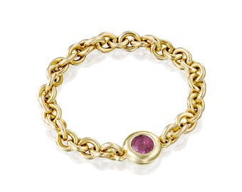 May include: A gold chain ring with a pink gemstone set in a bezel. The ring features a thick chain design with a circular pink stone in the center. The ring is made of gold and is set against a white background.