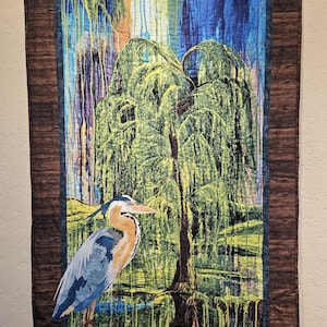 May include: A rectangular fabric wall hanging featuring a quilted scene of a great blue heron standing on a rock. The background depicts a weeping willow tree with green foliage against a blue and yellow sky, all framed by a dark brown border.