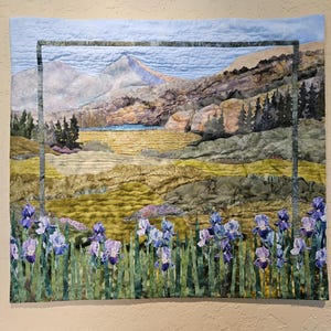 Può includere: Un paesaggio trapuntato con una catena montuosa, un lago e iris. La trapunta utilizza varie trame e colori di tessuto, tra cui blu, verdi e gialli. Una cornice scura circonda il paesaggio.