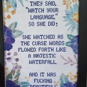 May include: A framed cross-stitch with a white background and blue and green floral accents. The text reads "They said, "Watch your language," so she did; She watched as the curse words flowed forth like a majestic waterfall. And it was fucking beautiful!"