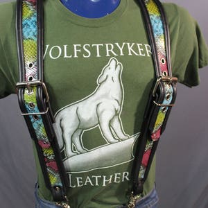 Peut inclure: Bretelles en cuir noir avec un motif de peau de serpent coloré. Les bretelles sont portées sur un t-shirt vert avec le texte "Wolfstryker Leather" imprimé sur le devant.