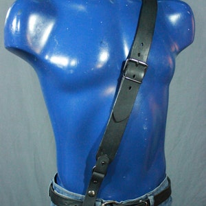 May include: Black leather shoulder strap with a silver buckle, worn over a blue mannequin torso. The strap is paired with a black leather belt and blue jeans. The mannequin is painted a glossy blue.