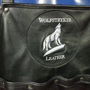 May include: Black leather apron with the embroidered logo of a howling wolf on a patch that says "WOLFSTRYKER LEATHER". The apron has pockets and is secured with silver rivets.