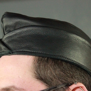May include: A black leather cap with a rounded crown and a small, angled brim. The cap is made of a textured leather material and is worn on the head. The cap is a classic style, often associated with military or aviation attire.
