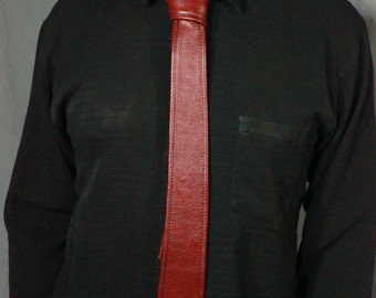 Peut inclure: Une cravate en cuir bordeaux se détache sur une chemise noire à manches longues. La cravate est de couleur unie et a un aspect texturé. La chemise a une poche sur le côté gauche. Le fond est gris uni.