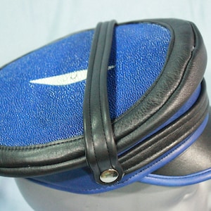 May include: A blue and black leather cap with a white stripe and a black leather strap. The cap has a rounded crown and a flat brim.