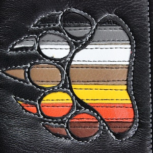 Puede incluir: Una cartera de cuero negro con un diseño de pata de oso colorido. La pata está hecha de tiras de cuero de diferentes colores cosidas juntas en un patrón de arcoíris.