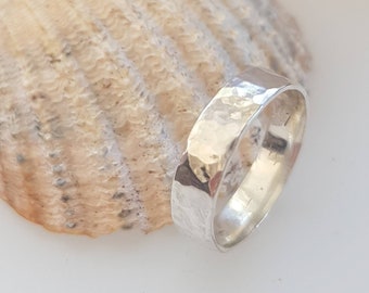 May include: A silver hammered band ring with a smooth, polished interior. The ring is displayed on a seashell.