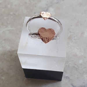 以下が含まれることがあります： ハンマーで叩いたローズゴールドのハートのアクセントが付いたシルバーリング2つ。リングは重ねられています。