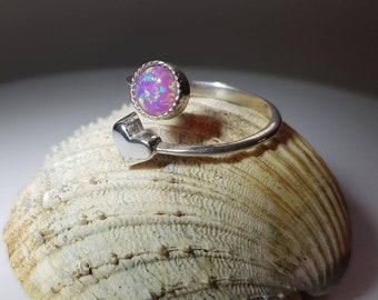 May include: A silver ring with a pink opal stone and a heart-shaped design. The ring is sitting on a seashell.
