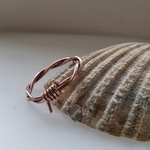 May include: A copper wire ring with a barbed wire design. The ring is resting on a seashell.