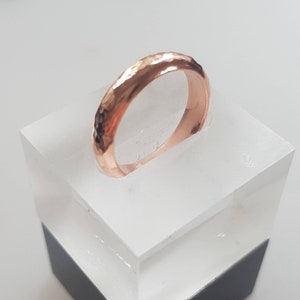 May include: A rose gold hammered band ring displayed on a white acrylic stand.