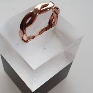 May include: A rose gold ring with a braided design. The ring is displayed on a white and black acrylic stand.