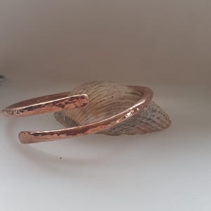 May include: A hammered copper bangle bracelet with a simple, curved design. The bracelet is shown resting on a piece of driftwood.