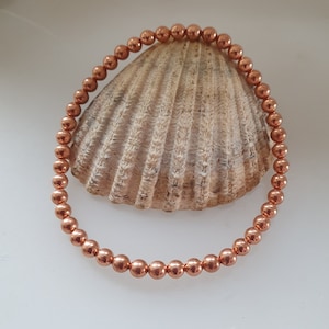 May include: A copper beaded bracelet with a shiny finish. The bracelet is made of small, round beads that are strung together on a thin cord. The bracelet is lying on a seashell.