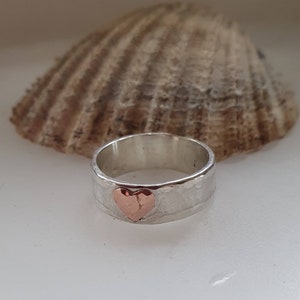 Puede incluir: Un anillo de banda de plata con textura martillada y un corazón de cobre en el centro.