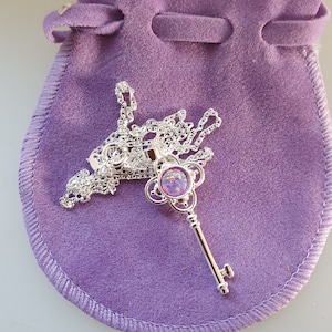 Puede incluir: Un collar con colgante de llave plateada con una piedra morada similar al ópalo en el centro, sobre una bolsa de terciopelo morado. El colgante de llave presenta detalles florales y una cadena delicada.