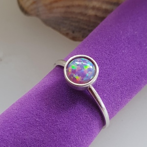 May include: A silver ring featuring a round, iridescent opal gemstone. The opal has a purple base with flashes of green, pink, and blue.