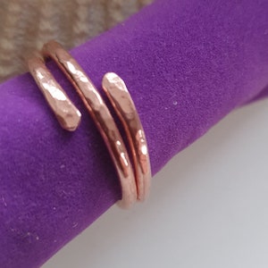 May include: A copper-coloured ring with a hammered texture. The ring has a wrap-around design with two parallel bands and a third band that curves around the finger. The ring is displayed on a purple fabric.