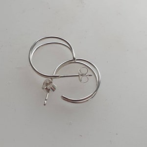 Può includere: Un paio di orecchini a cerchio argentati. Ogni orecchino presenta un design circolare a doppio strato. Gli orecchini hanno una chiusura a perno e farfalla. I gioielli sono esposti su una superficie bianca.