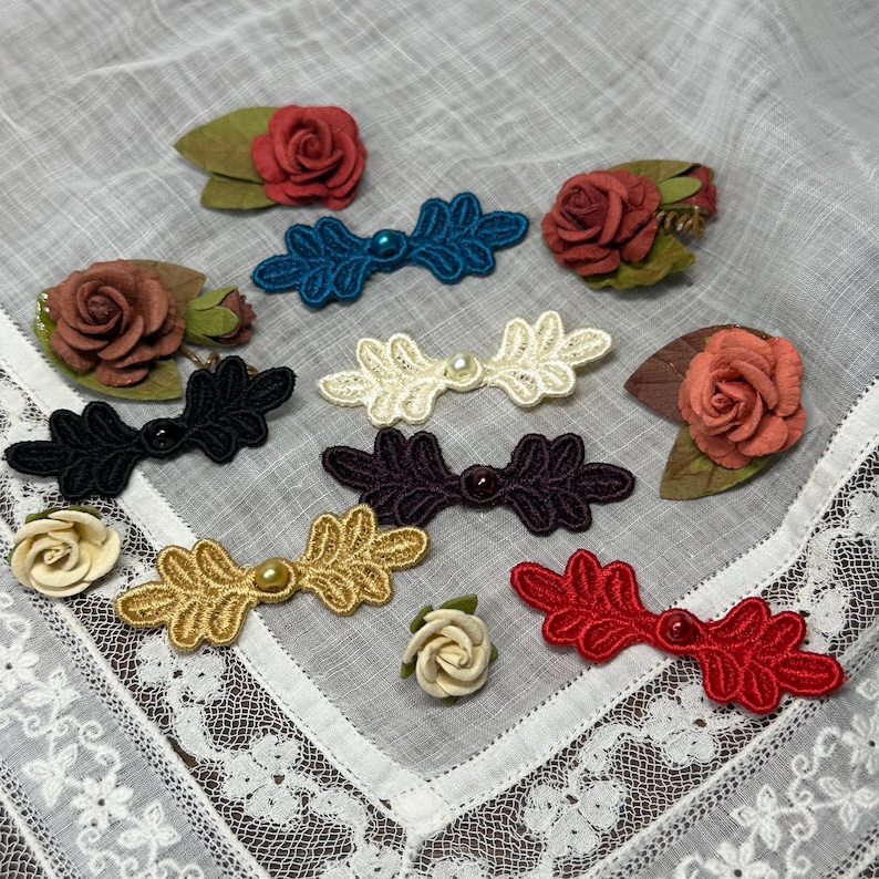 May include: A collection of decorative hair accessories and embellishments. The image features various colors of lace bows with pearl accents, along with small fabric roses in shades of red, brown, and cream. The items are arranged on a white, lacy surface.