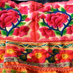 May include: Close-up of a colorful embroidered fabric with pink flowers and green leaves. The fabric has a fringe border and a multi-colored trim.