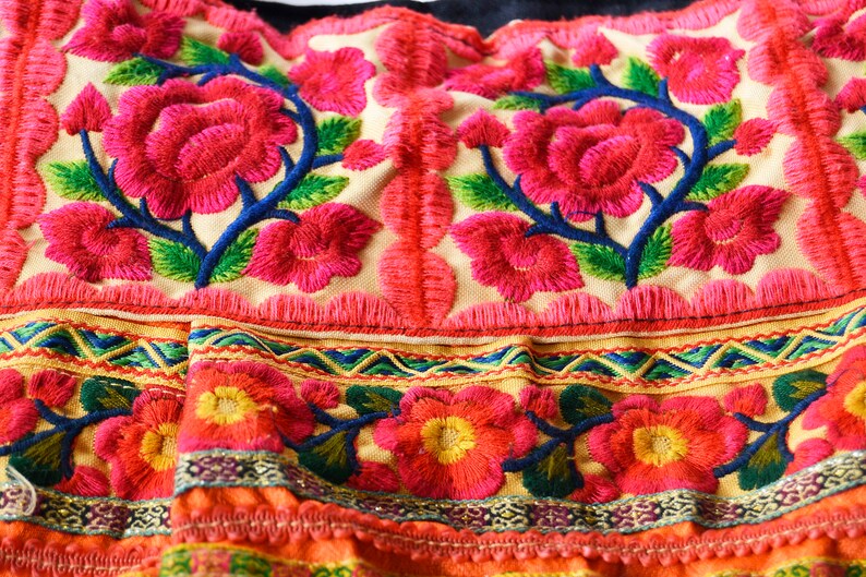 May include: Close-up of a colorful embroidered fabric with pink flowers and green leaves. The fabric has a fringe border and a multi-colored trim.
