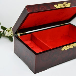 Peut inclure: Une bo&icirc;te &agrave; bijoux rectangulaire ouverte, avec une finition rouge fonc&eacute; brillante et un int&eacute;rieur en velours rouge. La bo&icirc;te a une quincaillerie de couleur laiton et un couvercle &agrave; charni&egrave;re. Une petite branche de fleurs blanches et de feuilles vertes est en arri&egrave;re-plan.