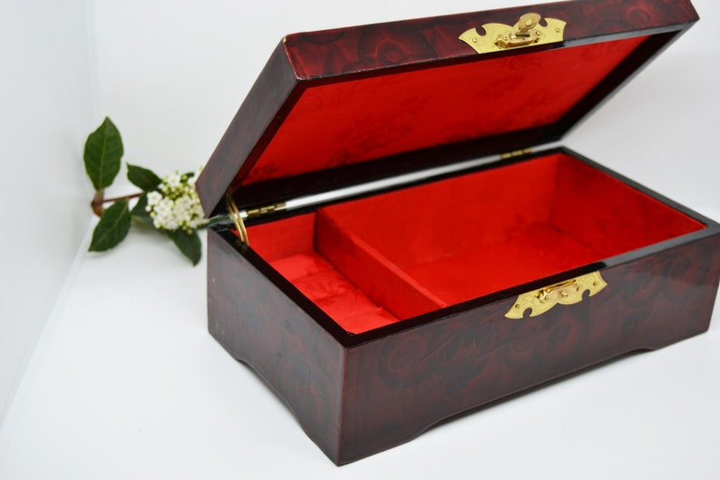 Peut inclure: Une bo&icirc;te &agrave; bijoux rectangulaire ouverte, avec une finition rouge fonc&eacute; brillante et un int&eacute;rieur en velours rouge. La bo&icirc;te a une quincaillerie de couleur laiton et un couvercle &agrave; charni&egrave;re. Une petite branche de fleurs blanches et de feuilles vertes est en arri&egrave;re-plan.