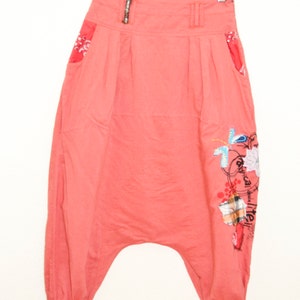 Può includere: Pantaloni color corallo con vita alta e vestibilità ampia. I pantaloni hanno una stampa a bandana rossa e bianca sulla cintura e sulle tasche. I pantaloni hanno una stampa grafica sulla gamba destra con il testo "tropical detail".