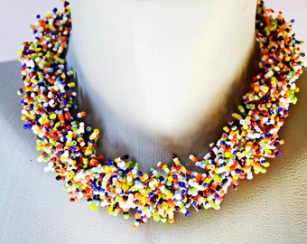 May include: A colorful beaded necklace made of small, multi-colored glass beads. The necklace is arranged in a cascading style, creating a layered and textured look.