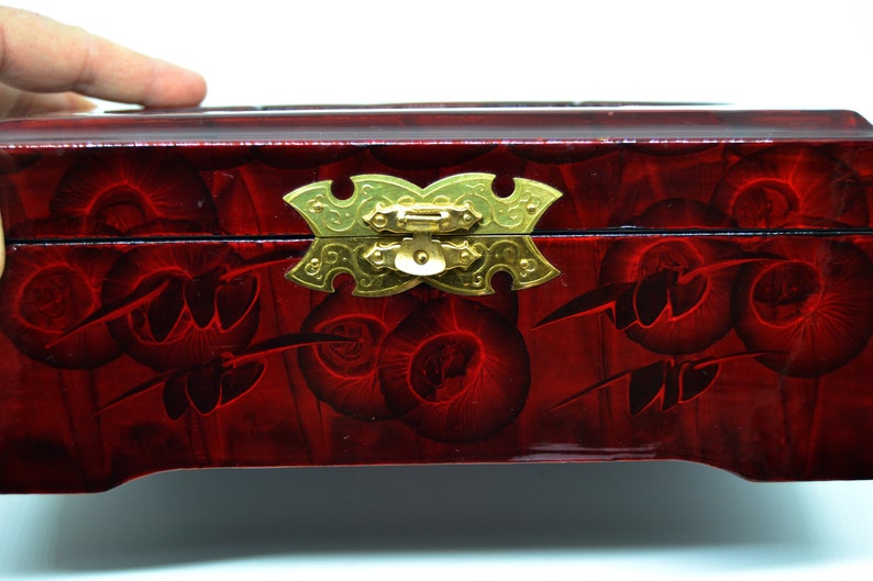 Peut inclure: Une bo&icirc;te en bois laqu&eacute; rouge avec un motif floral d&eacute;coratif. La bo&icirc;te est dot&eacute;e d'un fermoir et de charni&egrave;res dor&eacute;s. La surface a une finition brillante et le motif est de couleur rouge fonc&eacute;.