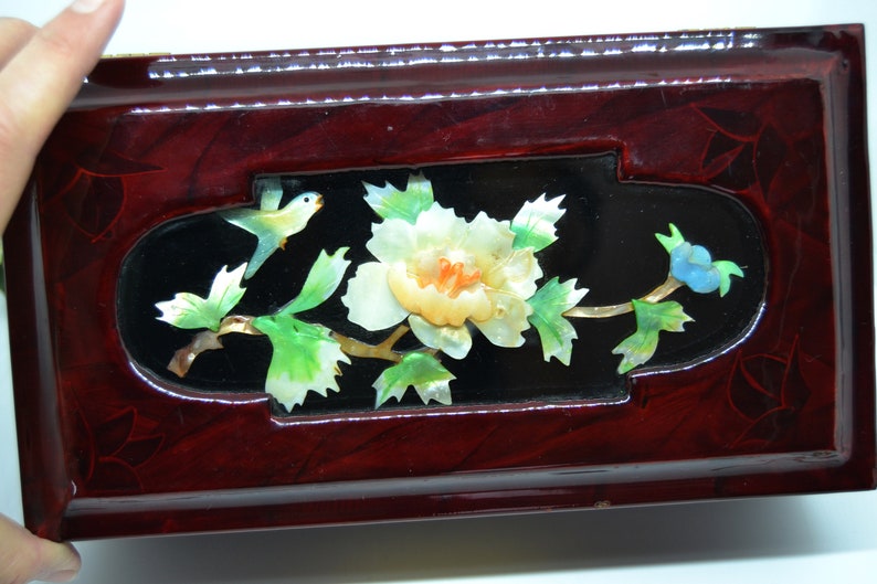 Peut inclure: Bo&icirc;te rectangulaire en bois laqu&eacute;, finition rouge fonc&eacute;. Le couvercle pr&eacute;sente une incrustation noire avec un motif floral et d'oiseaux en nacre. Le motif comprend une fleur blanche, des feuilles vertes et un oiseau bleu.