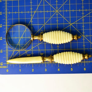 May include: A set of two vintage magnifying glass and letter opener tools with ivory-colored handles and gold accents. The tools are on a blue cutting mat with inch markings.