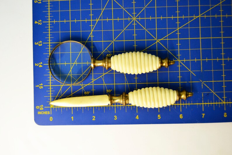 May include: A set of two vintage magnifying glass and letter opener tools with ivory-colored handles and gold accents. The tools are on a blue cutting mat with inch markings.