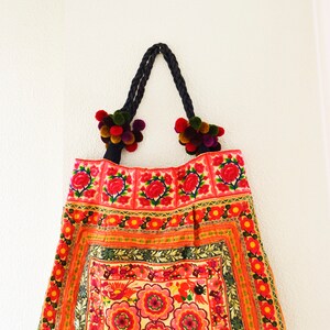 May include: A red and pink floral embroidered tote bag with black braided handles and colorful pom-poms.