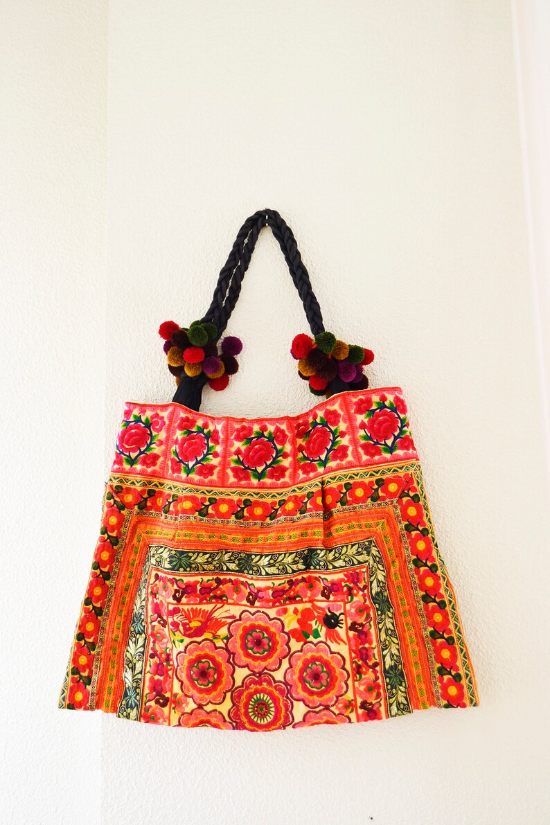 May include: A red and pink floral embroidered tote bag with black braided handles and colorful pom-poms.