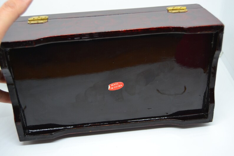 Peut inclure: Une bo&icirc;te rectangulaire en bois laqu&eacute; avec une finition brillante brun-rouge fonc&eacute;. La bo&icirc;te a un couvercle &agrave; charni&egrave;re avec une quincaillerie de couleur laiton. Un autocollant rouge avec le texte blanc "MADE IN CHINA" est appos&eacute; au fond.