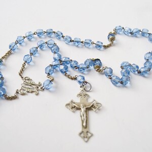 May include: A blue glass rosary with a silver crucifix and a silver charm. The rosary beads are faceted and have a light blue color.