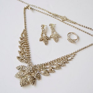 Pu&ograve; includere: Un set di gioielli color argento con una collana, orecchini e un anello. La collana presenta un grande design floreale con strass. Gli orecchini sono orecchini pendenti con un design floreale simile. L'anello &egrave; una semplice fascia con strass.