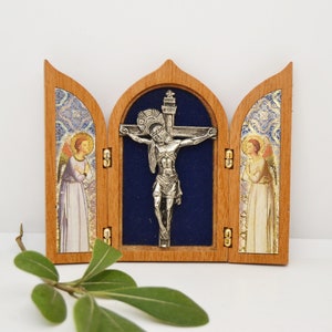 May include: A wooden triptych with a silver crucifix in the center panel. The side panels feature images of angels with gold accents. The triptych is closed and has a blue velvet interior.
