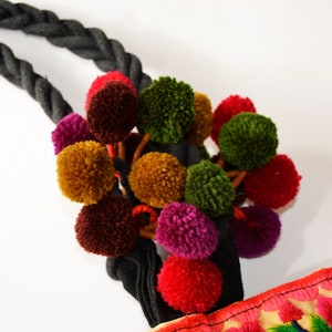 May include: A black rope with a cluster of colorful pom-poms attached. The pom-poms are in shades of red, green, brown, and purple. The rope is tied to a fabric bag with embroidered flowers.