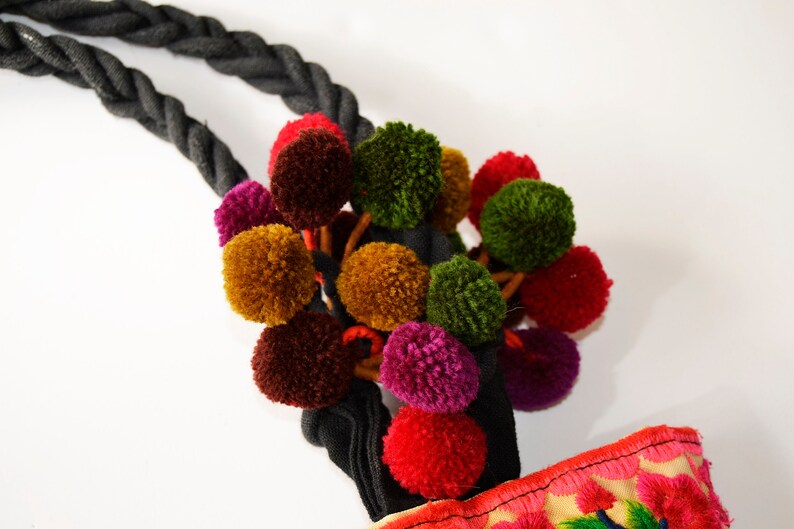 May include: A black rope with a cluster of colorful pom-poms attached. The pom-poms are in shades of red, green, brown, and purple. The rope is tied to a fabric bag with embroidered flowers.