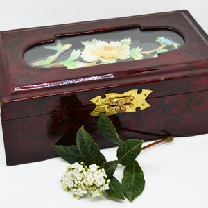 Peut inclure: Une bo&icirc;te rectangulaire en bois laqu&eacute; de couleur bordeaux fonc&eacute;, orn&eacute;e d'un motif floral d&eacute;coratif. Le couvercle comporte une fen&ecirc;tre ovale transparente avec un motif floral peint. Un fermoir dor&eacute; s&eacute;curise la bo&icirc;te. Une branche de fleurs blanches et de feuilles vertes est &agrave; proximit&eacute;.