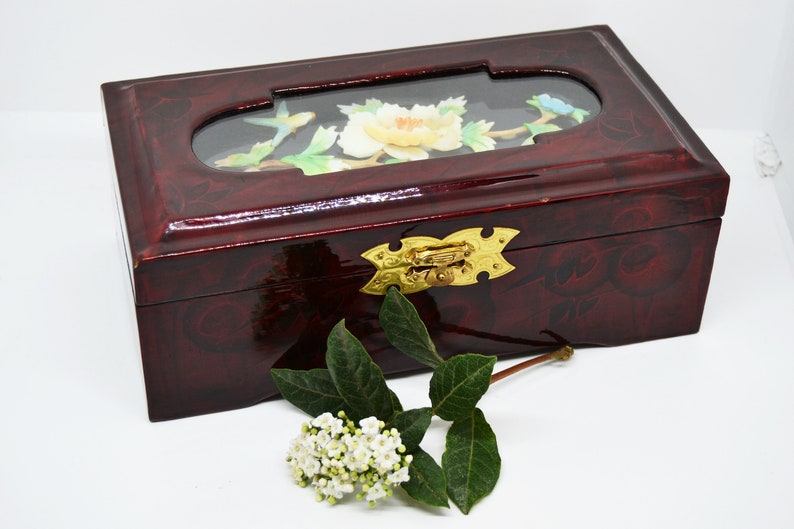 Peut inclure: Une bo&icirc;te rectangulaire en bois laqu&eacute; de couleur bordeaux fonc&eacute;, orn&eacute;e d'un motif floral d&eacute;coratif. Le couvercle comporte une fen&ecirc;tre ovale transparente avec un motif floral peint. Un fermoir dor&eacute; s&eacute;curise la bo&icirc;te. Une branche de fleurs blanches et de feuilles vertes est &agrave; proximit&eacute;.