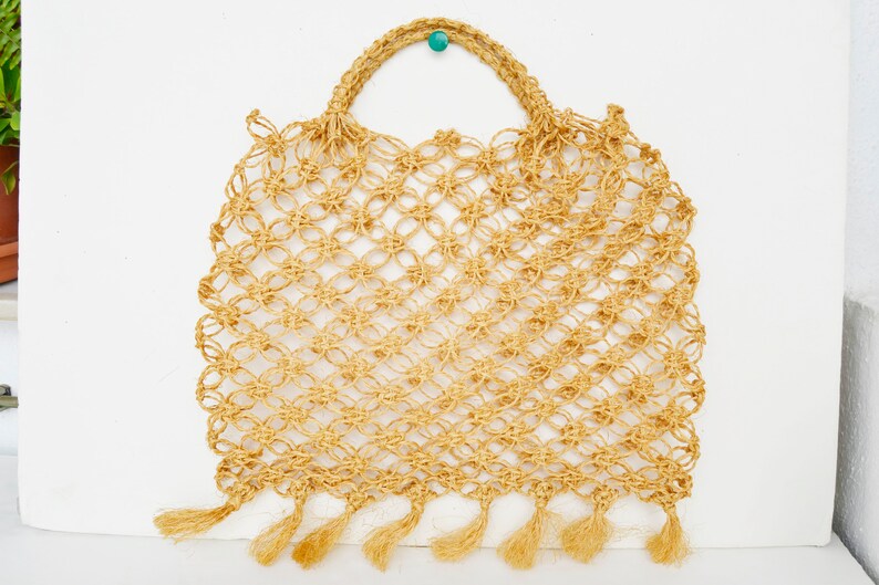 May include: A beige crocheted tote bag with fringe detailing. The bag has a knotted design and a turquoise bead on the handle.