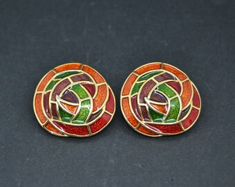 May include: A pair of round, clip-on earrings with a stained-glass design. The earrings feature a gold-tone metal frame and are filled with red, orange, green, and brown enamel. The earrings have a spiral pattern.