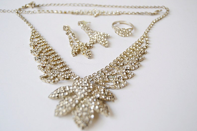 Pu&ograve; includere: Un set di gioielli color argento con una collana, orecchini e un anello. La collana presenta una catena delicata con un grande ciondolo a forma di foglia adornato con scintillanti strass. Anche gli orecchini sono adornati con strass e pendono da piccoli cerchi argentati. L'anello &egrave; una semplice fascia con un piccolo design a forma di foglia.